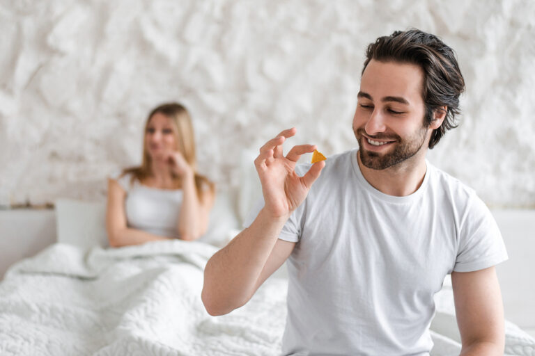 Smiling man holding a Tadalafil Enhanced Gummy, used for improving erectile dysfunction, while a woman in the background looks away, sitting on a bed.